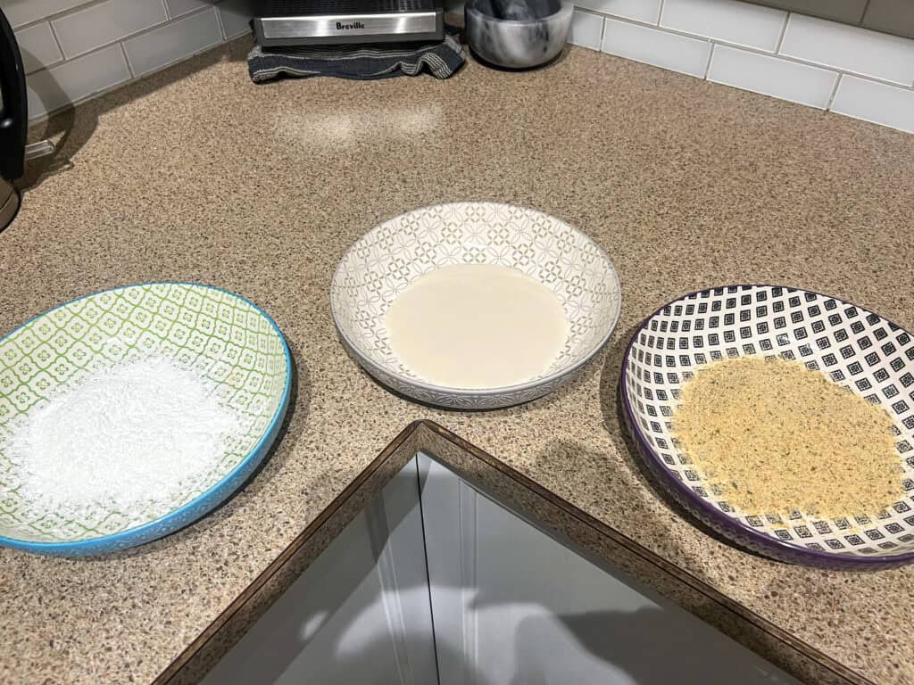 Coating ingredients in decorative bowls on a kitchen countertop for cooking preparation.